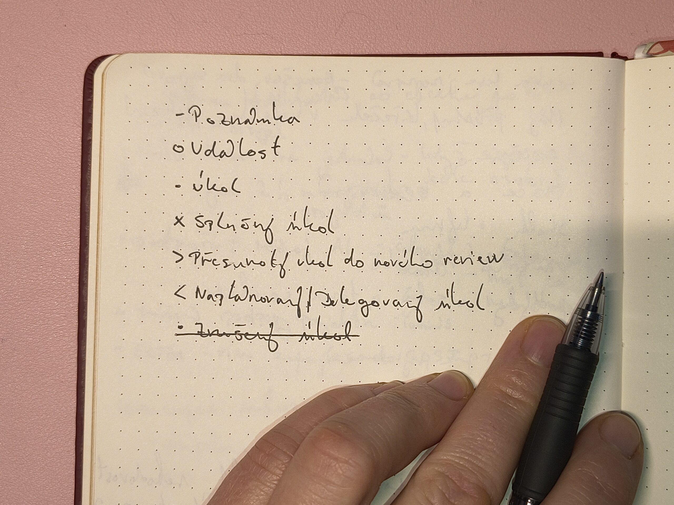 Detail ruky s gelovým perem nad otevřeným tečkovaným sešitem, ukazující jednoduchý systém značek pro plánování metodou Bullet Journal jako alternativu k digitálním aplikacím.